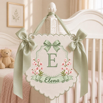 May include: A cream-colored scalloped wall hanging featuring a large green letter E, the name Elena in cursive script, and pink floral accents. The sign is decorated with a green ribbon bow at the top and two matching side bows with long ribbons.