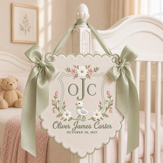 May include: A cream-colored fabric wall hanging with scalloped edges, featuring embroidered floral motifs, a central duckling, the initials oJC, and the text Oliver James Carter OCTOBER 20, 2027. It is adorned with two sage green satin ribbons.