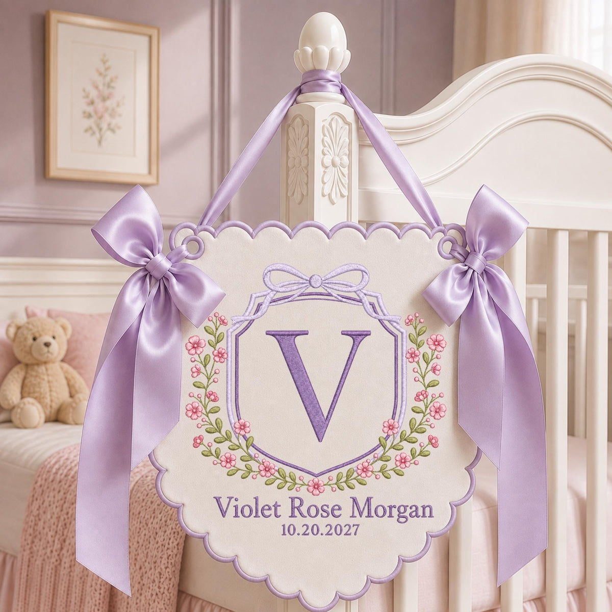 May include: A cream-colored scalloped wall hanging featuring an embroidered purple letter V inside a floral wreath. The text reads Violet Rose Morgan and 10.20.2027. Two lavender satin ribbons are tied in bows on the top corners for hanging.