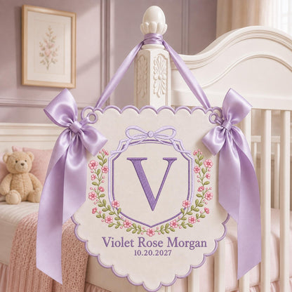 May include: A cream-colored scalloped wall hanging featuring an embroidered purple letter V inside a floral wreath. The text reads Violet Rose Morgan and 10.20.2027. Two lavender satin ribbons are tied in bows on the top corners for hanging.