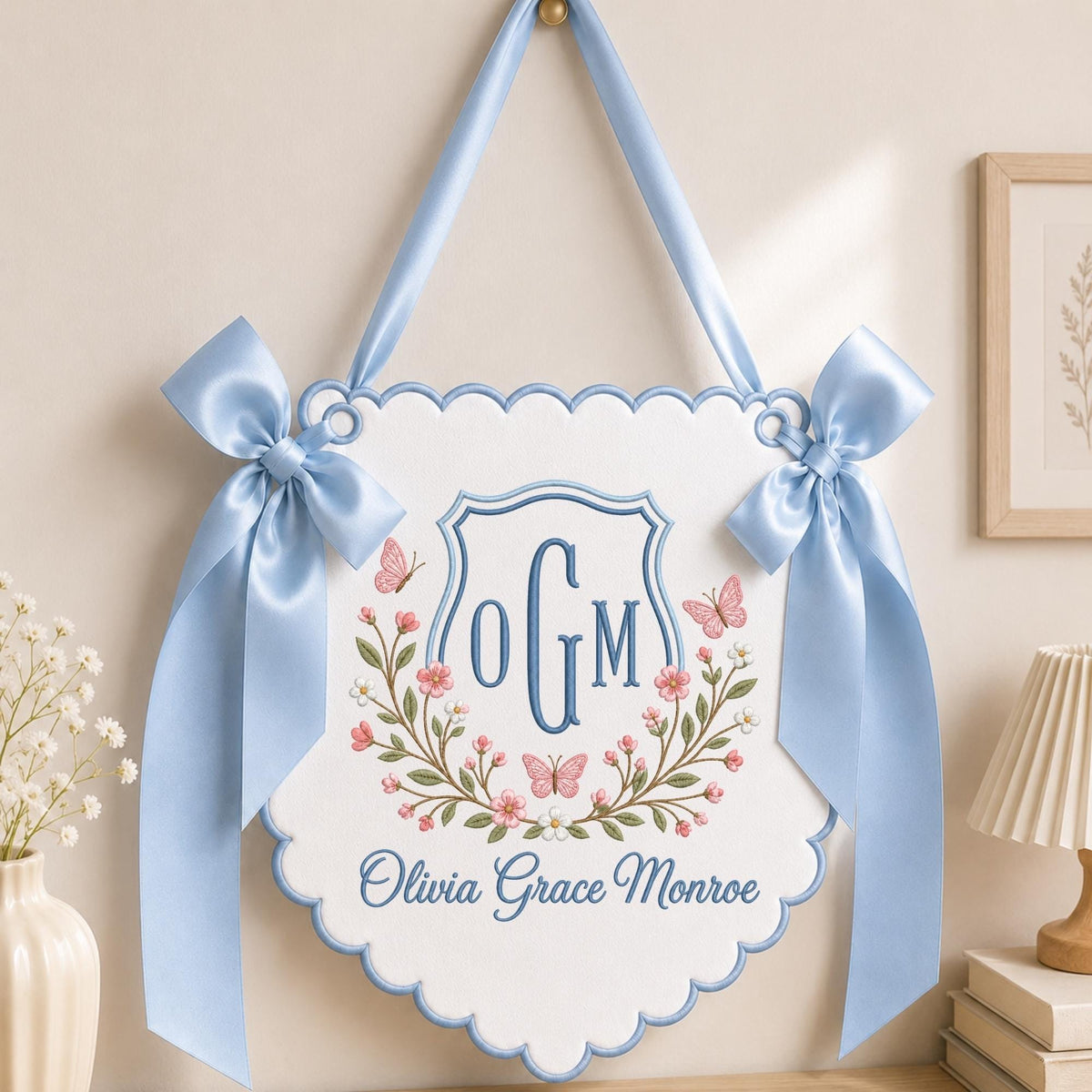 May include: A white scalloped wall hanging featuring a blue embroidered shield with the monogram OGM, surrounded by pink floral vines and butterflies. The name Olivia Grace Monroe is embroidered below in blue, with light blue satin ribbon bows.