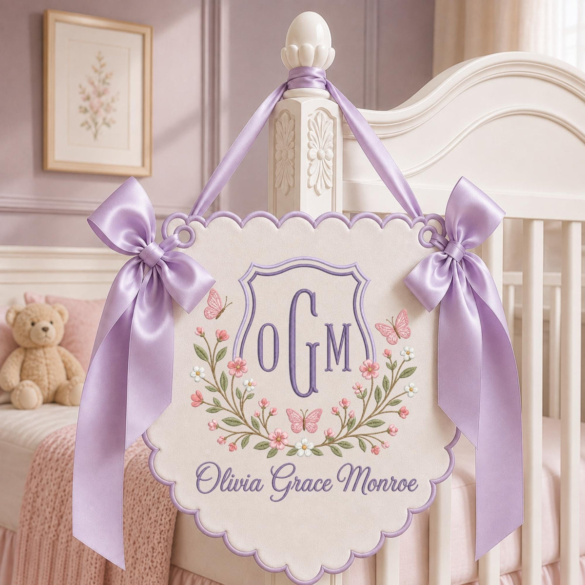 May include: A cream-colored scalloped wall hanging featuring an embroidered purple monogram OGM, floral wreath, and butterflies. The sign displays the name Olivia Grace Monroe in purple script and is suspended by two lavender satin ribbons.