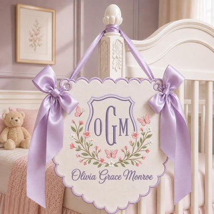 May include: A cream-colored scalloped wall hanging featuring an embroidered purple monogram OGM, floral wreath, and butterflies. The sign displays the name Olivia Grace Monroe in purple script and is suspended by two lavender satin ribbons.