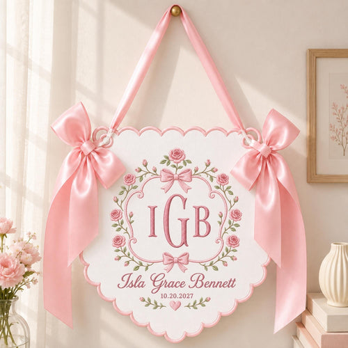 May include: A white scalloped wall hanging featuring pink embroidered roses, a central monogram with the letters IGB, and the text Isla Grace Bennett 10.20.2027. The piece is suspended by a pink satin ribbon and adorned with two large pink satin bows.