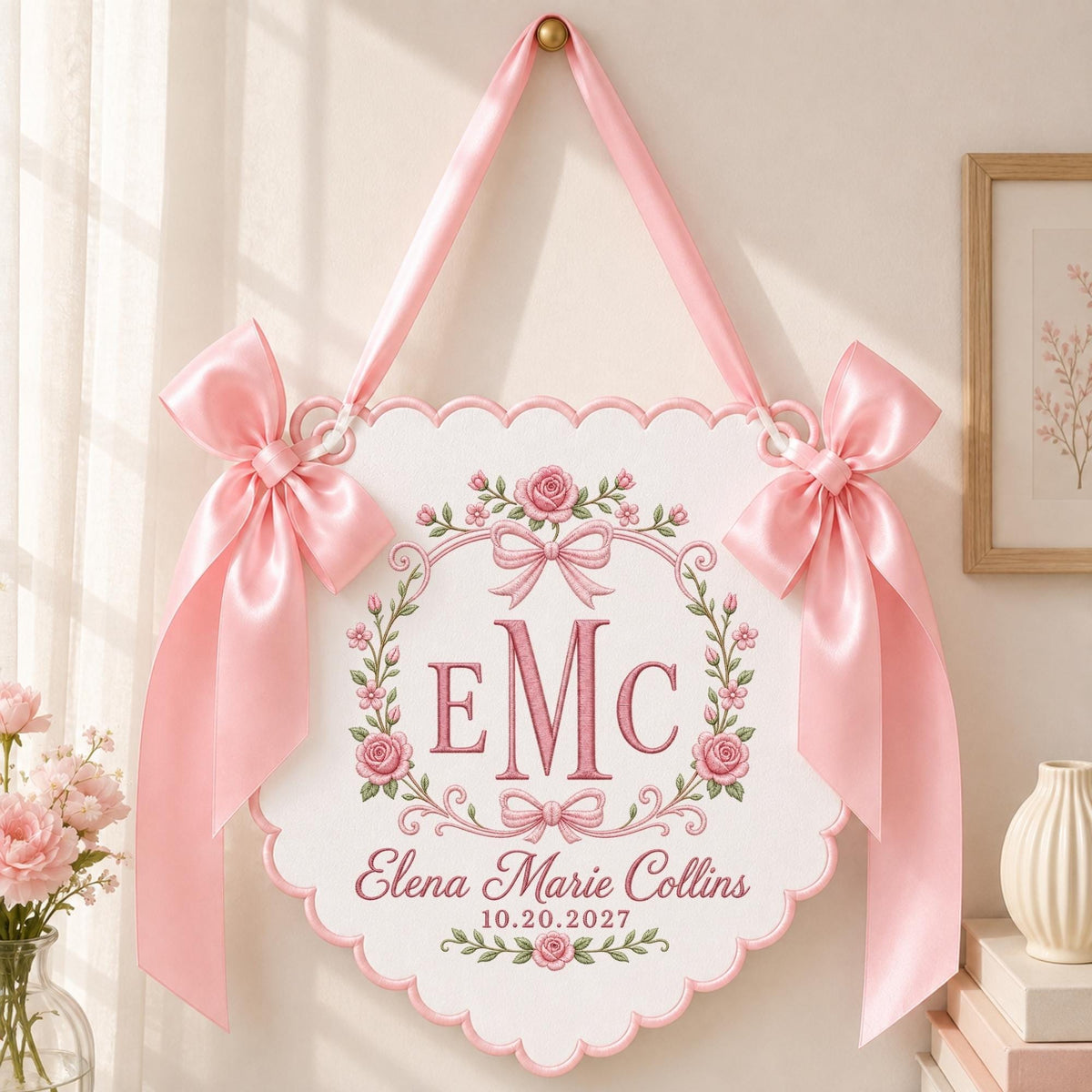 May include: A white scalloped wall hanging featuring a pink embroidered floral wreath, the monogram "EMC", and the text "Elena Marie Collins 10.20.2027". The sign is suspended by a pink satin ribbon and decorated with two large pink satin bows on each side.