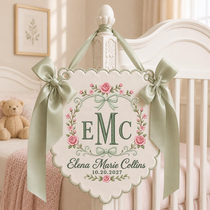 May include: A cream-colored scalloped wall hanging featuring a sage green ribbon bow at the top and sides. The center displays the monogram EMC in sage green, surrounded by a floral wreath of pink roses and green leaves, with the text Elena Marie Collins 10.20.2027.