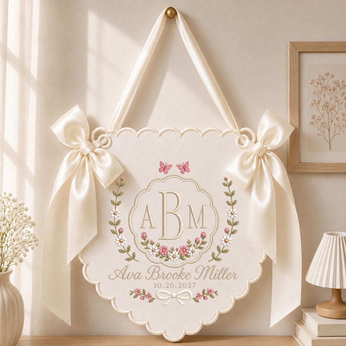 May include: A cream-colored fabric wall hanging with scalloped edges, featuring two satin bows, embroidered floral wreaths, and two butterflies. The center displays the monogram ABM, the name Ava Brooke Miller, and the date 10.20.2027 in gold thread.