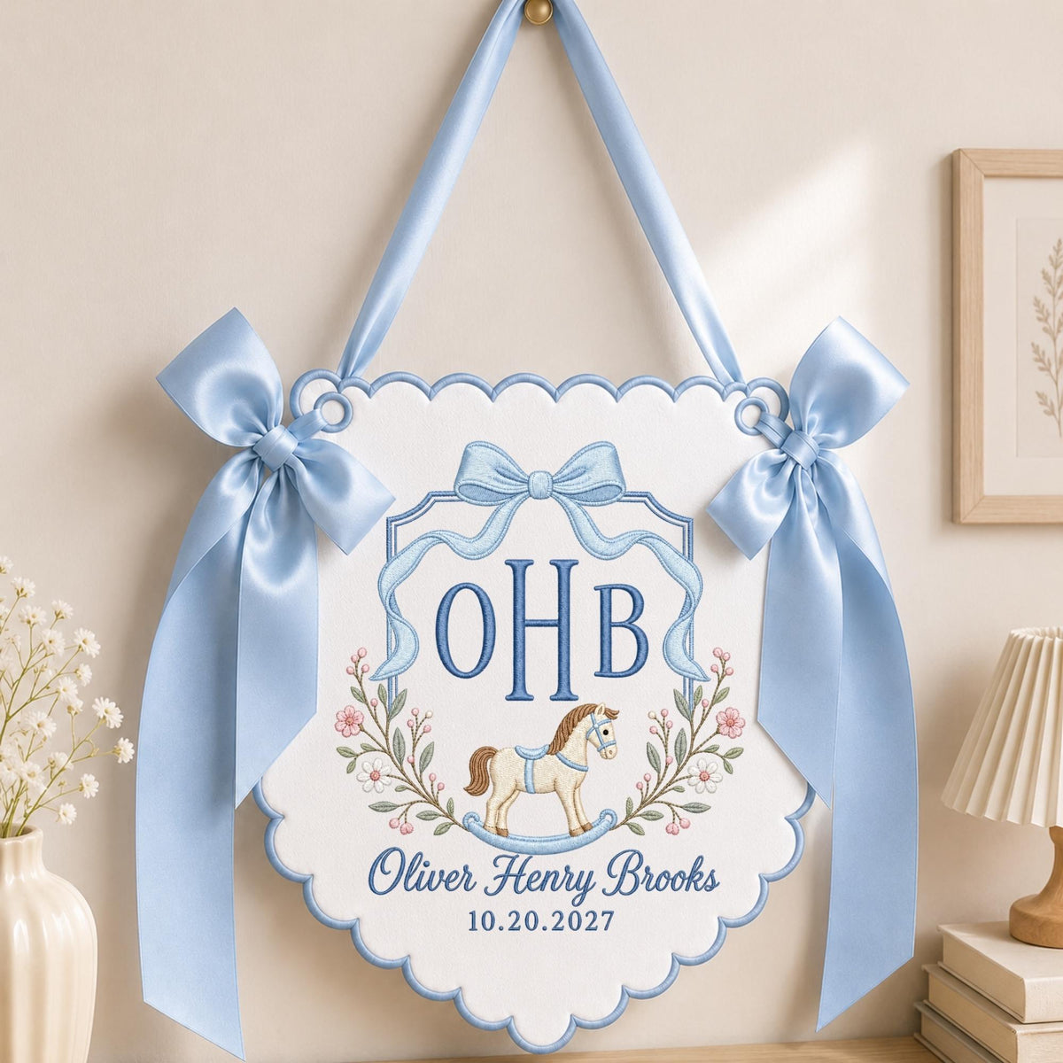 May include: A white scalloped fabric wall hanging featuring light blue satin ribbon bows on each side and a matching hanging loop. The embroidery displays the initials OHB, a rocking horse, and the text Oliver Henry Brooks 10.20.2027.