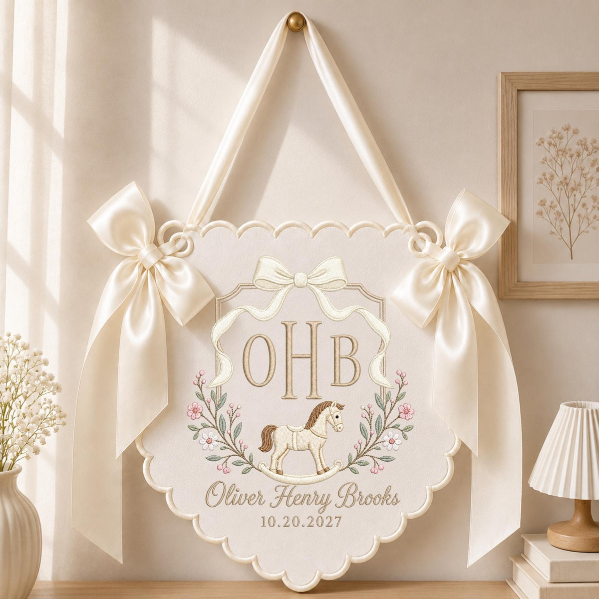 May include: A cream-colored fabric wall hanging with scalloped edges, featuring two large satin bows on the sides. The center displays the embroidered initials OHB, a rocking horse illustration, floral vines, and the text Oliver Henry Brooks 10.20.2027.