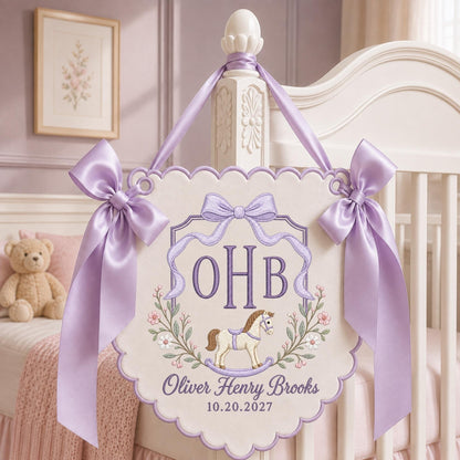 May include: A cream-colored fabric wall hanging with scalloped edges, featuring lavender embroidery of the initials OHB, a rocking horse, and floral accents. It includes the text Oliver Henry Brooks and 10.20.2027, finished with lavender satin ribbons.