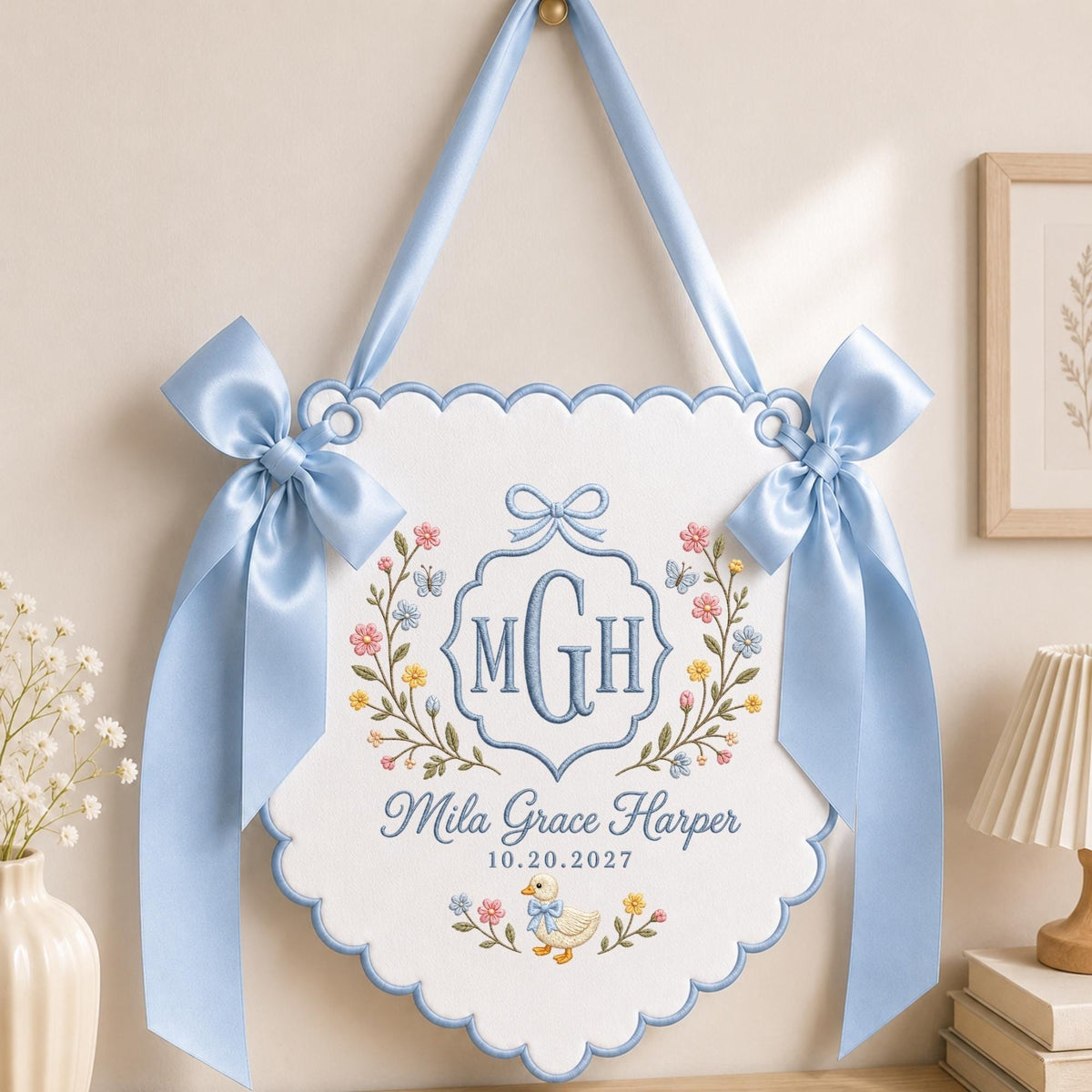 May include: A white scalloped wall hanging with light blue satin ribbon bows and trim. It features embroidered floral vines, a central monogram MGH, the name Mila Grace Harper, the date 10.20.2027, and a small duck illustration in blue and gold.