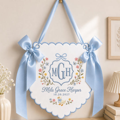 May include: A white scalloped wall hanging with light blue satin ribbon bows and trim. It features embroidered floral vines, a central monogram MGH, the name Mila Grace Harper, the date 10.20.2027, and a small duck illustration in blue and gold.