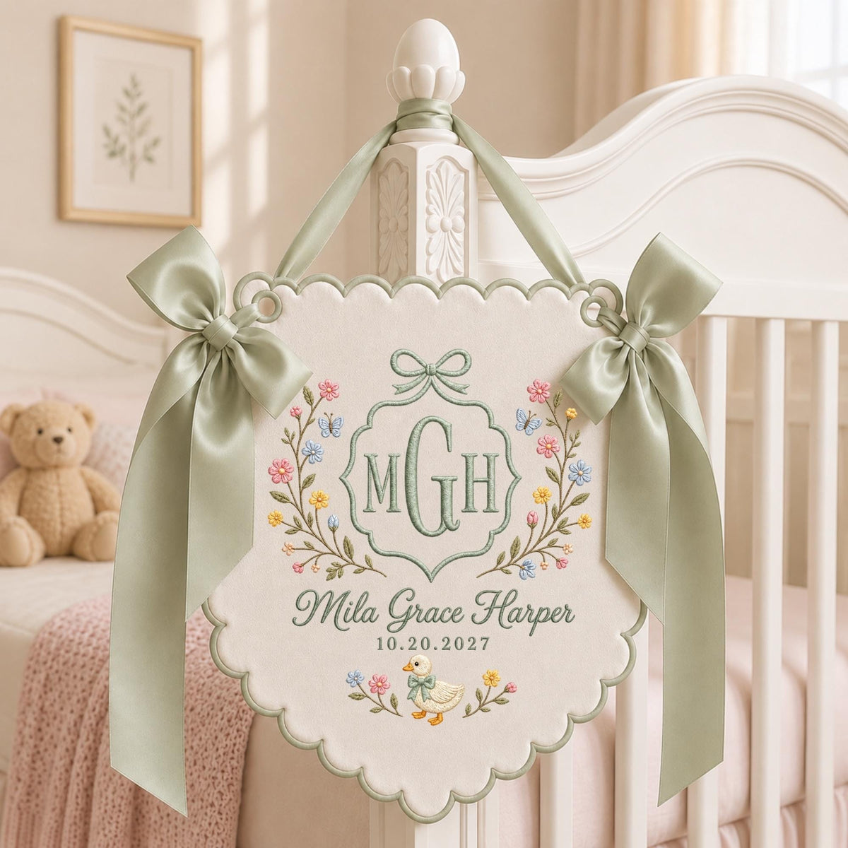 May include: A cream-colored fabric wall hanging with scalloped edges and sage green ribbon bows. It features embroidered floral vines, a central monogram MGH, the name Mila Grace Harper, the date 10.20.2027, and a small duck illustration.