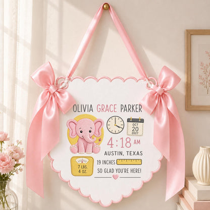 May include: A white scalloped wall hanging featuring a pink elephant illustration, the name Olivia Grace Parker, birth details including 4:18 AM, Oct 20 2027, Austin, Texas, 19 inches, and 7 lbs. 4 oz., accented by two large pink satin bows.