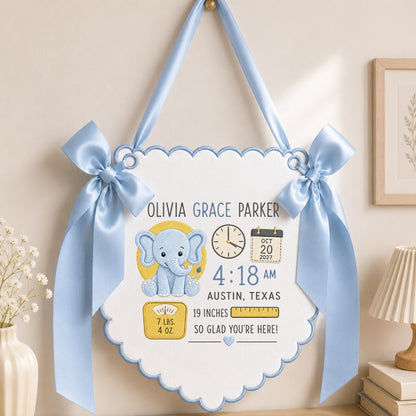 May include: A white scalloped wall hanging with light blue satin ribbon bows and trim. It features an embroidered blue elephant, a clock showing 4:18 AM, a calendar for Oct 20 2027, and text including Olivia Grace Parker, Austin, Texas, 7 lbs. 4 oz., and 19 inches.