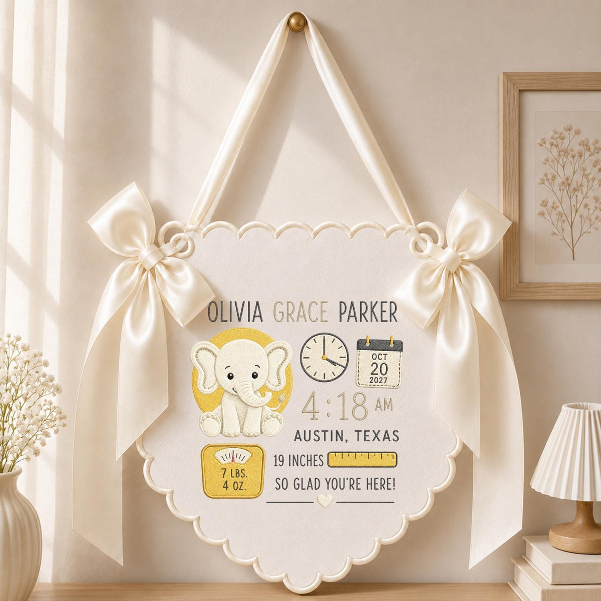 May include: A cream-colored scalloped wall hanging with satin ribbons and bows, featuring a cartoon elephant, a clock, a calendar showing Oct 20 2027, a scale reading 7 lbs. 4 oz., a ruler showing 19 inches, and the text Olivia Grace Parker Austin, Texas.