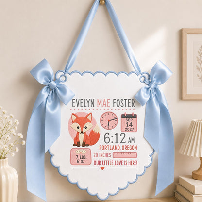 May include: A white scalloped wall hanging with light blue trim and satin bows, featuring a fox illustration and text: EVELYN MAE FOSTER, SEP 14 2027, 6:12 AM, PORTLAND, OREGON, 7 LBS. 6 OZ., 20 INCHES, and OUR LITTLE LOVE IS HERE! with a clock icon.