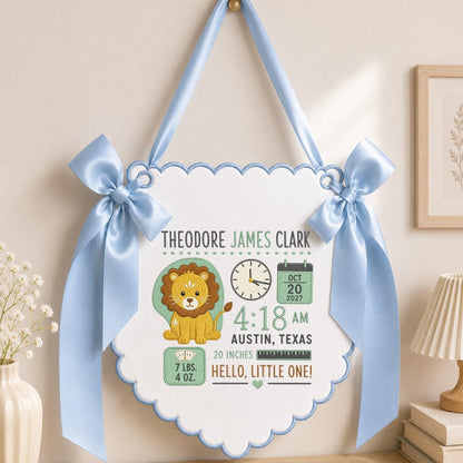 May include: A white scalloped wall hanging with light blue ribbon bows and trim. It features a cartoon lion, a clock showing 4:18 AM, a calendar for Oct 20 2027, and text: Theodore James Clark, Austin, Texas, 20 inches, 7 lbs. 4 oz., Hello, Little One!