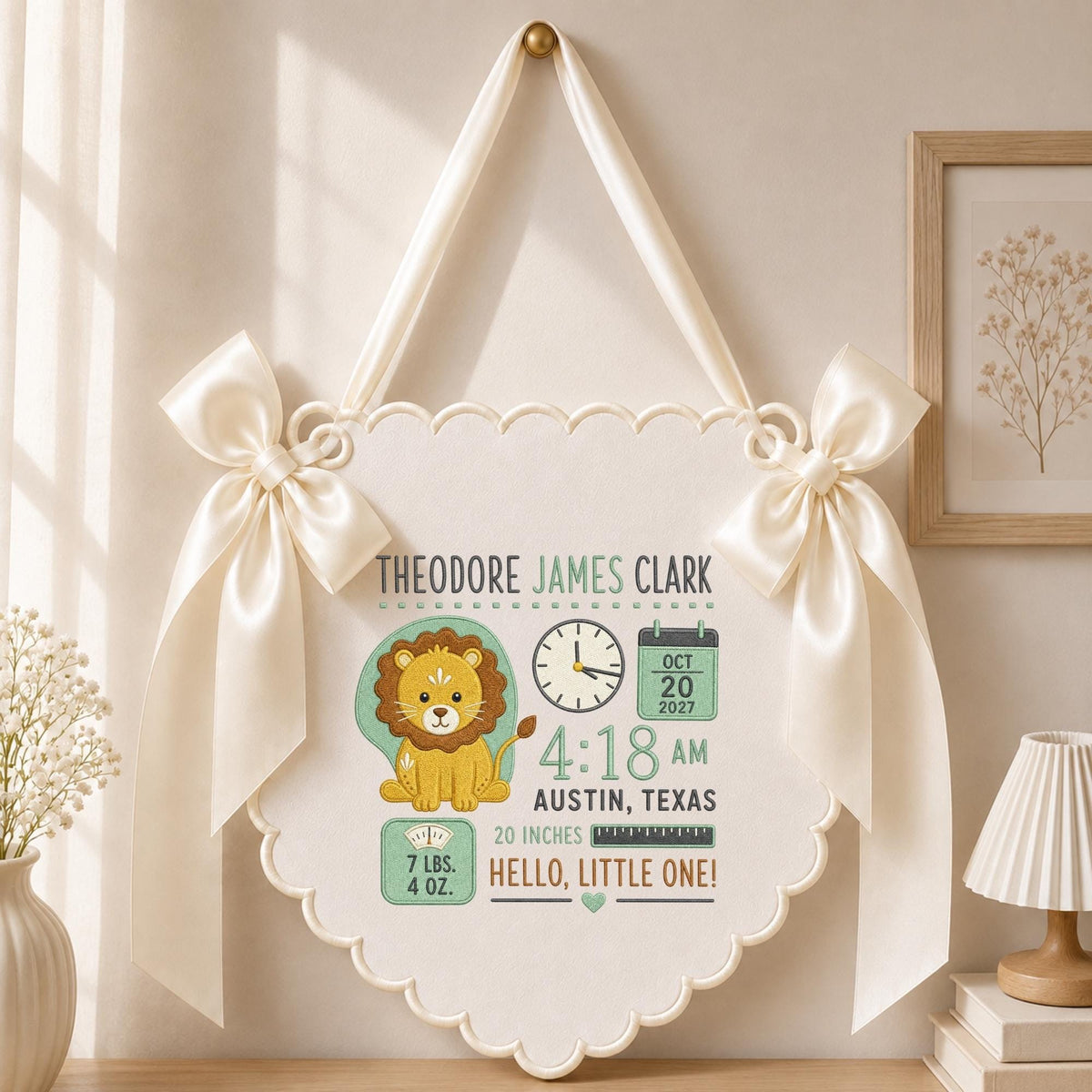 May include: A cream-colored scalloped wall hanging featuring a lion illustration, a clock, a calendar showing Oct 20 2027, and text including Theodore James Clark, 4:18 AM, Austin, Texas, 20 inches, 7 lbs. 4 oz., and Hello, Little One! with satin bows.