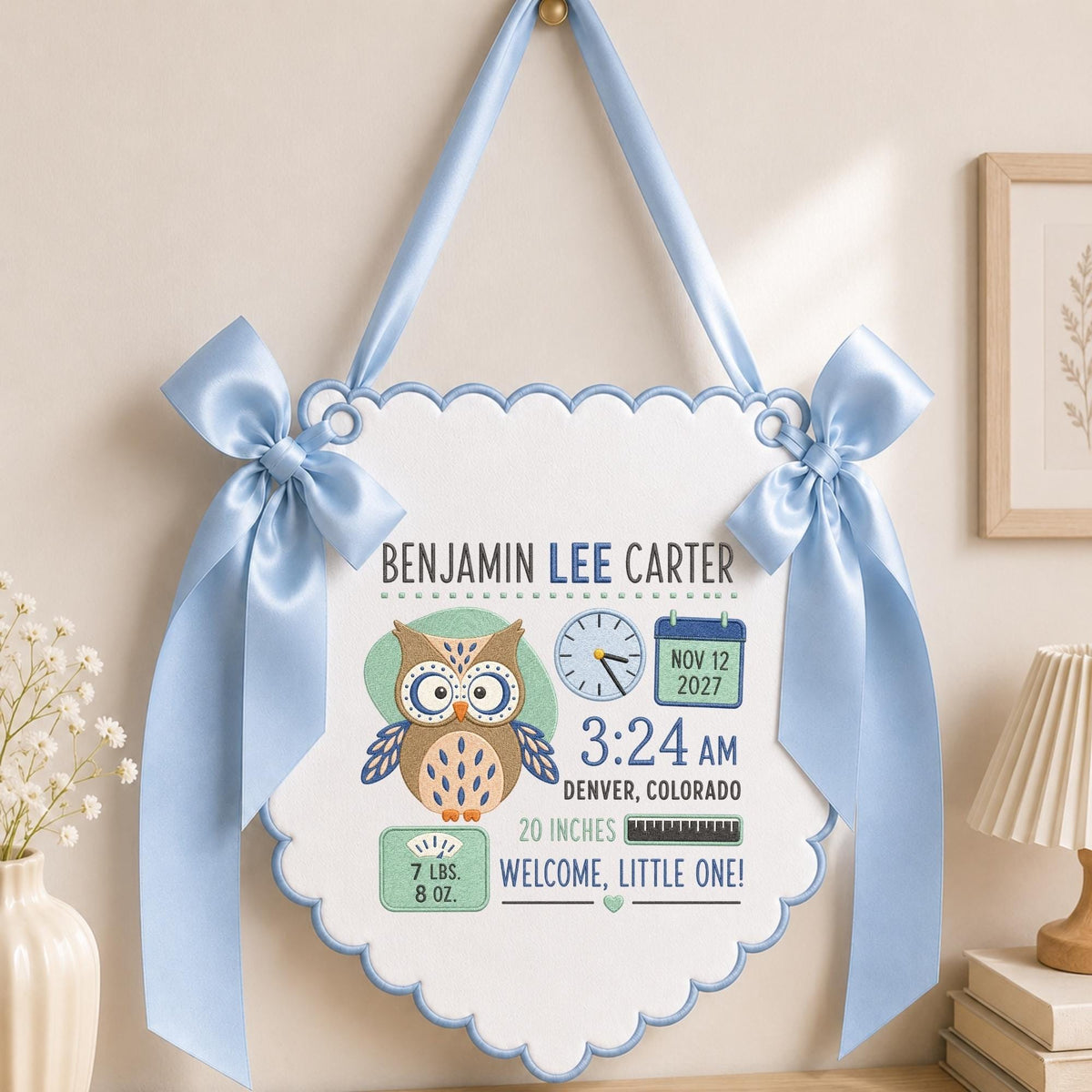 May include: A white scalloped wall hanging with light blue trim and satin bows, featuring an embroidered brown owl, a clock, a calendar showing Nov 12 2027, a scale reading 7 lbs. 8 oz., a ruler showing 20 inches, and the text Benjamin Lee Carter.