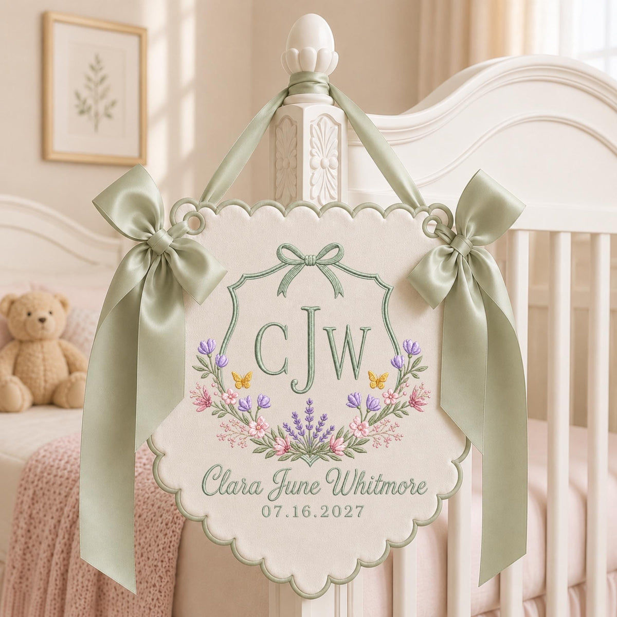 May include: A cream-colored fabric wall hanging with scalloped edges, featuring sage green satin ribbon bows. The embroidery includes the initials CJW, a floral wreath with lavender and pink flowers, and the text Clara June Whitmore 07.16.2027.