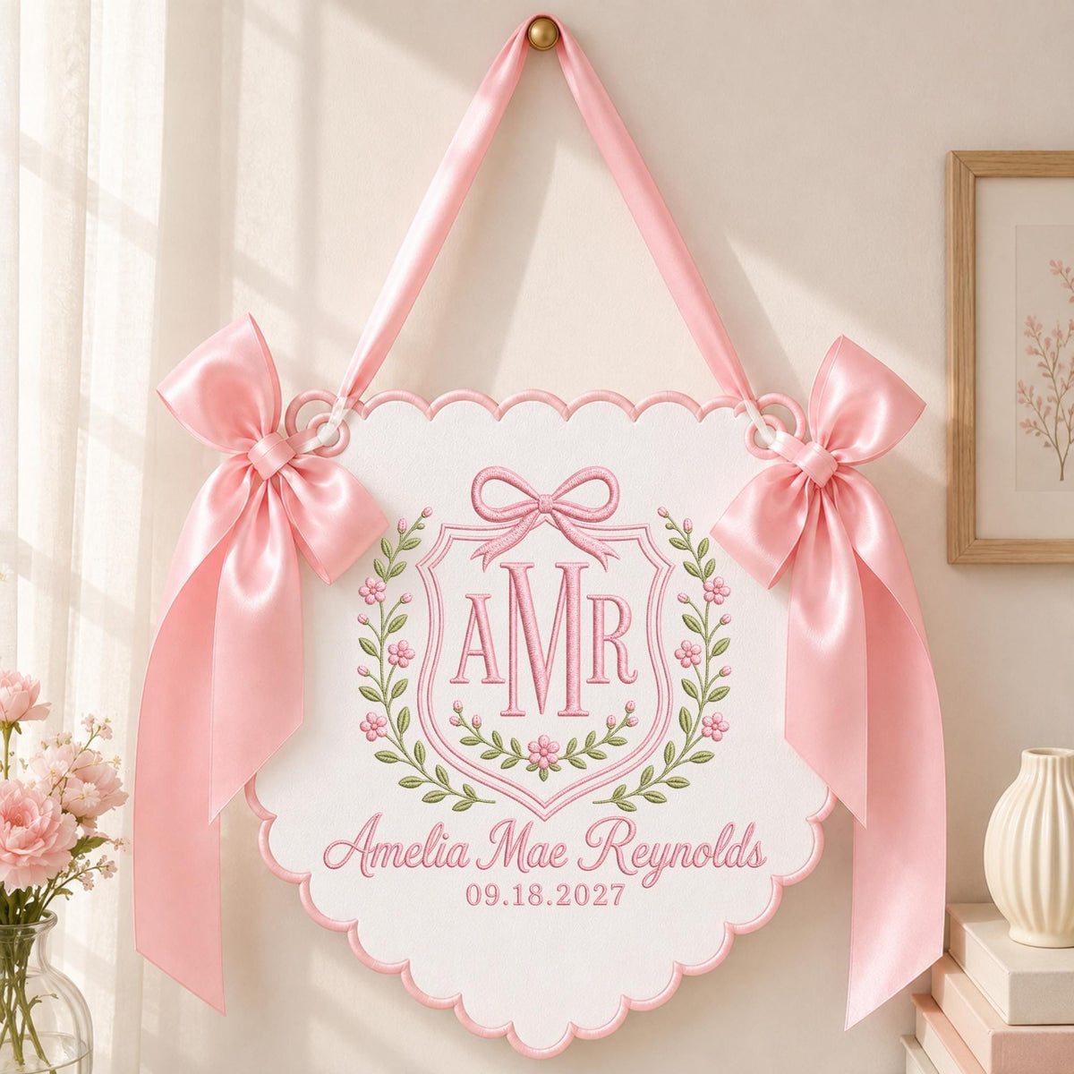 May include: A white scalloped wall hanging featuring pink satin ribbon bows on each side and a top hanging loop. The center displays an embroidered pink crest with the monogram AMR, surrounded by floral vines, the name Amelia Mae Reynolds, and 09.18.2027.
