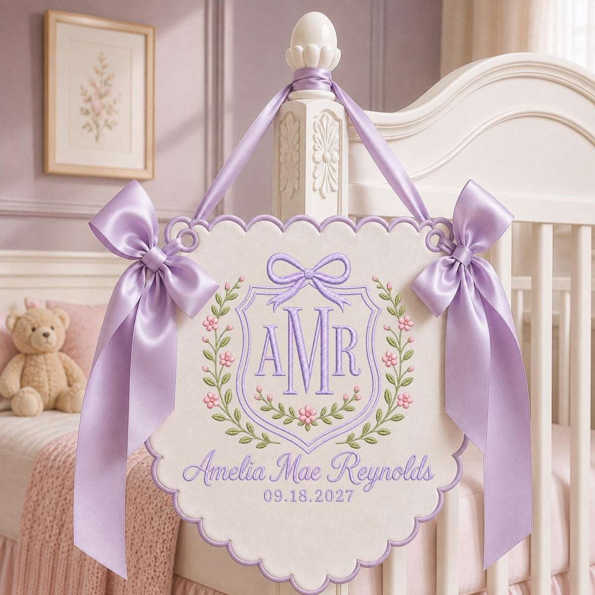 May include: A cream-colored scalloped wall hanging featuring lavender embroidery of the monogram AMR inside a shield, surrounded by floral vines. The text Amelia Mae Reynolds and 09.18.2027 is embroidered below. Two lavender satin bows adorn the top corners.