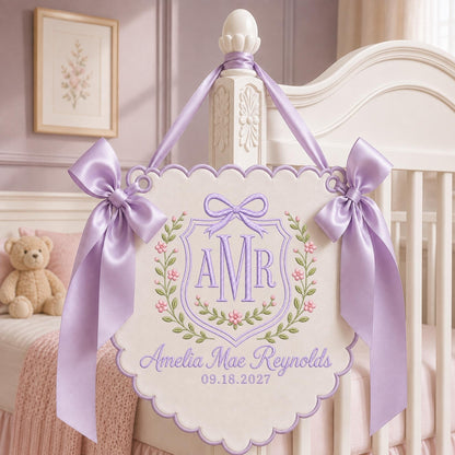 May include: A cream-colored scalloped wall hanging featuring lavender embroidery of the monogram AMR inside a shield, surrounded by floral vines. The text Amelia Mae Reynolds and 09.18.2027 is embroidered below. Two lavender satin bows adorn the top corners.