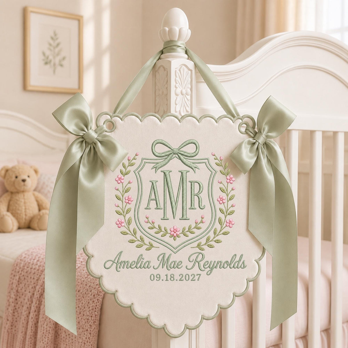May include: A cream-colored fabric wall hanging with scalloped edges, featuring sage green embroidery of the monogram AMR inside a floral crest. Below, the name Amelia Mae Reynolds and the date 09.18.2027 are embroidered. Two sage green satin bows adorn the top.