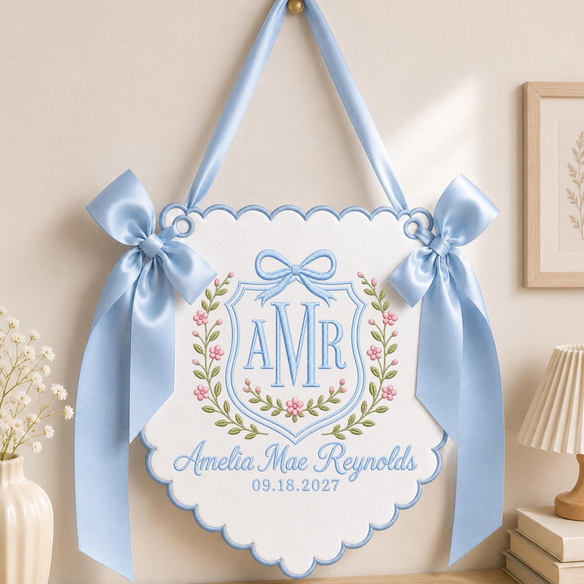 May include: A white scalloped wall hanging featuring light blue embroidery of the monogram AMR inside a shield, surrounded by a floral wreath. The text reads Amelia Mae Reynolds and 09.18.2027, accented by two large light blue satin bows.