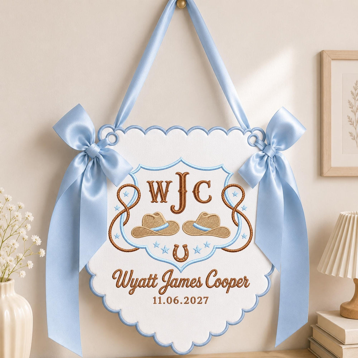 May include: A white scalloped wall hanging featuring light blue satin ribbon bows and trim. The embroidery includes the monogram WJC, two tan cowboy hats, a horseshoe, stars, and the text Wyatt James Cooper with the date 11.06.2027.