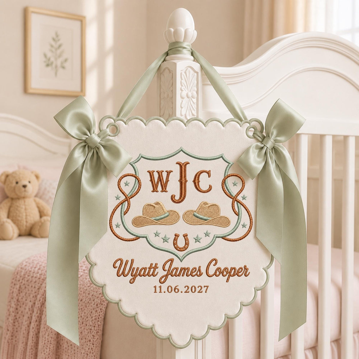 May include: A cream-colored fabric wall hanging with scalloped edges, featuring embroidered brown cowboy hats, a horseshoe, and the text WJC, Wyatt James Cooper, and 11.06.2027. It is adorned with two sage green satin ribbons tied in bows.