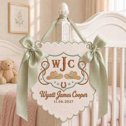 May include: A cream-colored fabric wall hanging with scalloped edges, featuring embroidered brown cowboy hats, a horseshoe, and the text WJC, Wyatt James Cooper, and 11.06.2027. It is adorned with two sage green satin ribbons tied in bows.