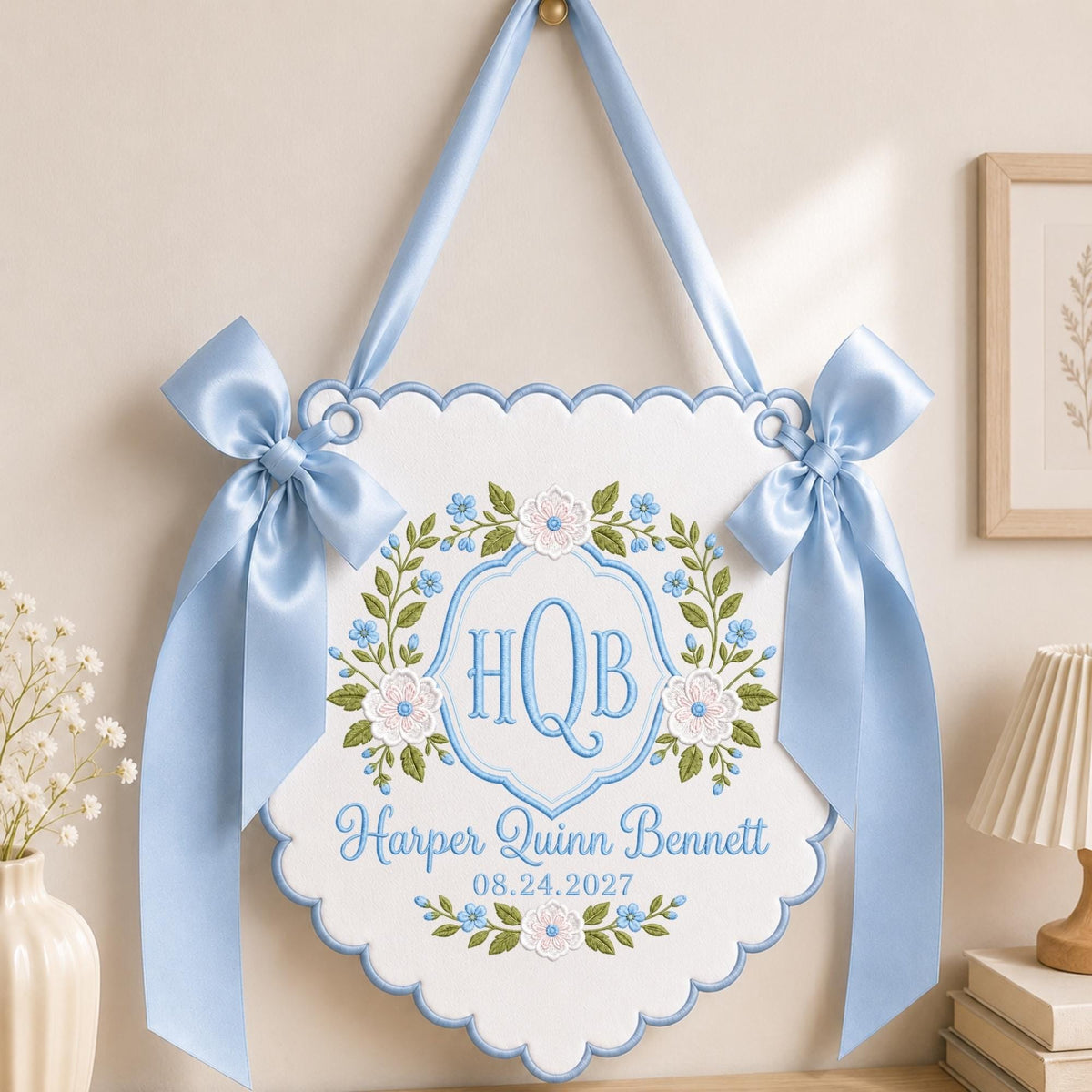 May include: A white scalloped wall hanging featuring light blue satin ribbon bows on each side. The center displays an embroidered floral wreath in green and blue surrounding the monogram HQB, with the name Harper Quinn Bennett and 08.24.2027.