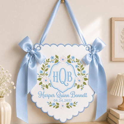 May include: A white scalloped wall hanging featuring light blue satin ribbon bows on each side. The center displays an embroidered floral wreath in green and blue surrounding the monogram HQB, with the name Harper Quinn Bennett and 08.24.2027.