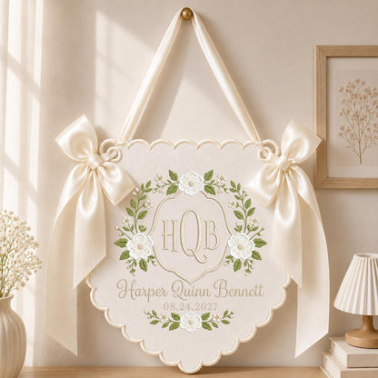 May include: A cream-colored scalloped wall hanging featuring an embroidered floral wreath surrounding the monogram HQB. Below the monogram, the name Harper Quinn Bennett and the date 08.24.2027 are embroidered. Two cream satin bows adorn the top corners.