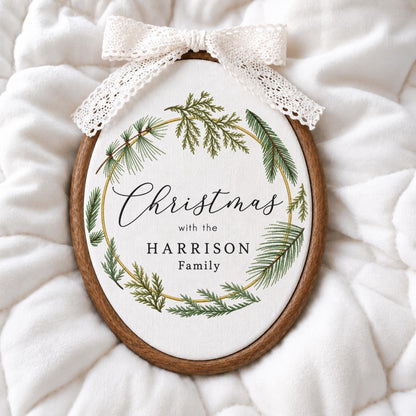 May include: An oval wooden embroidery hoop frame containing white fabric with a printed green pine needle wreath. The text reads "Christmas with the HARRISON Family". A white lace ribbon bow is attached to the top of the wooden frame.