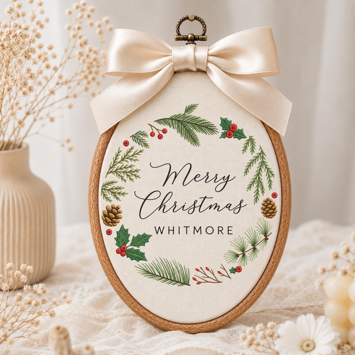 May include: An oval embroidery hoop frame containing off-white fabric with a printed wreath of green pine branches, red berries, and brown pinecones. The text Merry Christmas and WHITMORE is centered in black. A large cream satin bow sits on top.
