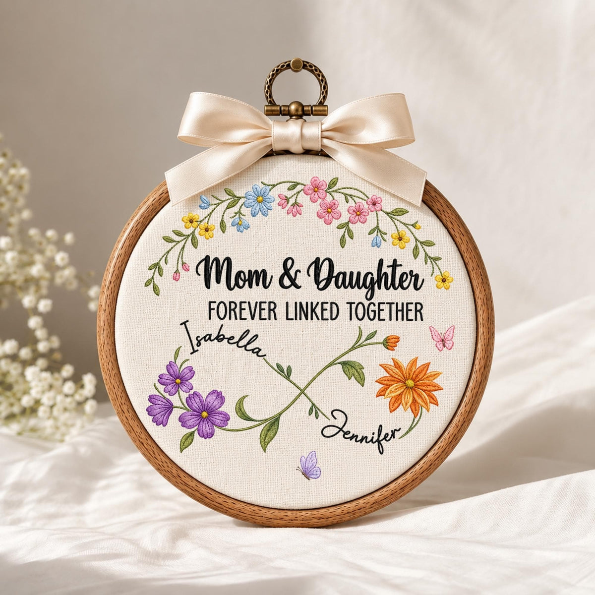 May include: A circular wooden embroidery hoop containing white fabric with the text Mom &amp; Daughter FOREVER LINKED TOGETHER. The design features a floral infinity symbol with the names Isabella and Jennifer, accented by colorful flowers and butterflies.