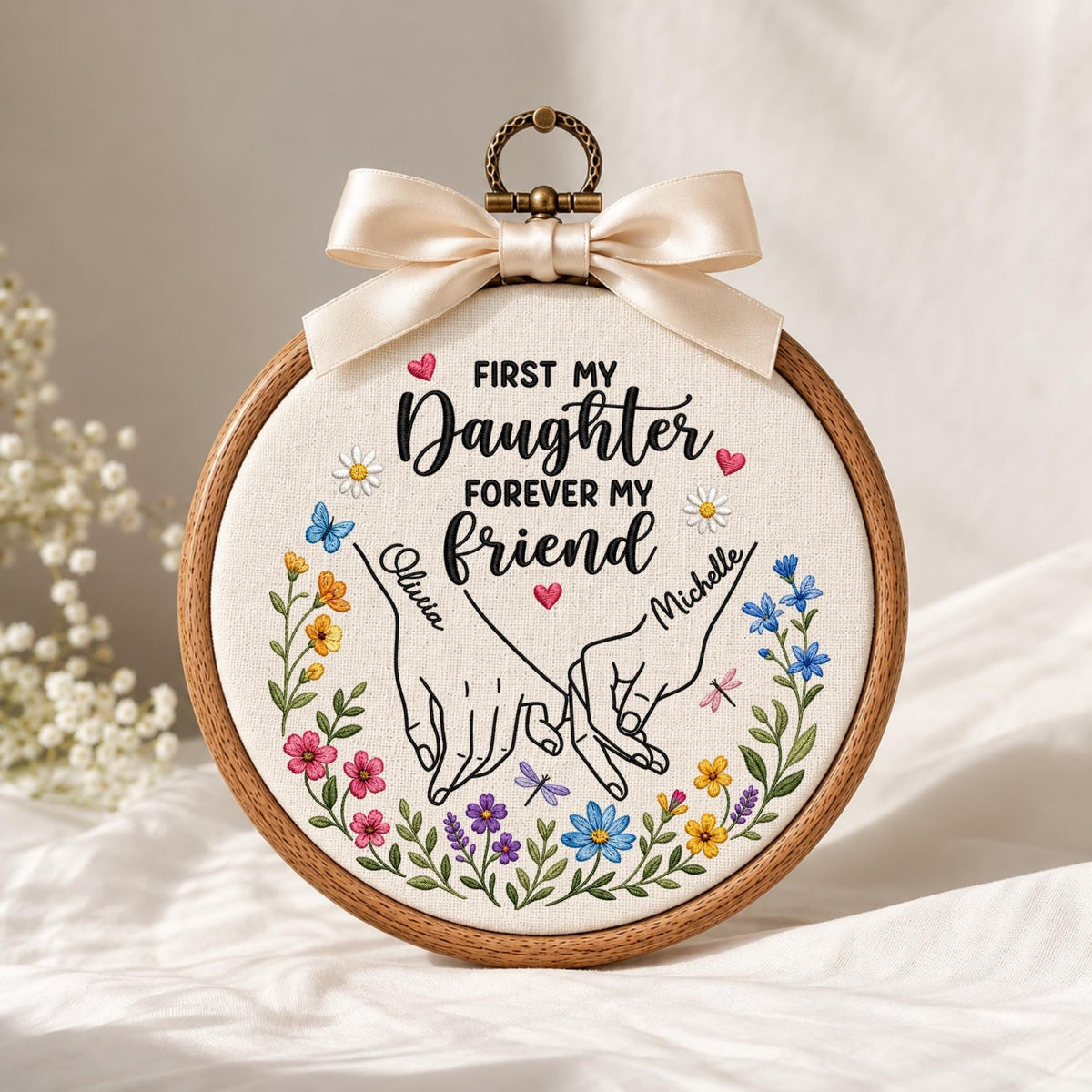 May include: A circular wooden embroidery hoop containing fabric with the embroidered text First My Daughter Forever My Friend, two hands holding pinky fingers, names Olivia and Michelle, and colorful floral accents, topped with a cream ribbon.