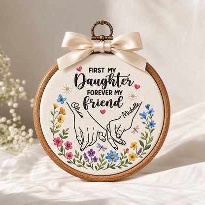 May include: A circular wooden embroidery hoop containing fabric with the embroidered text First My Daughter Forever My Friend, two hands holding pinky fingers, names Olivia and Michelle, and colorful floral accents, topped with a cream ribbon.