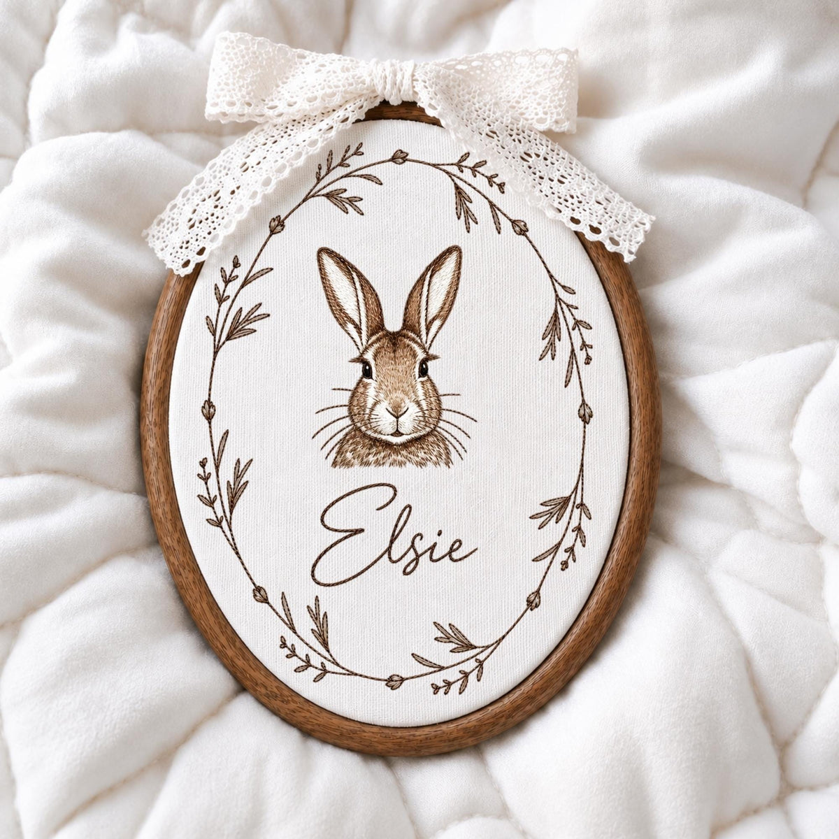 May include: An oval wooden embroidery hoop containing white fabric with a brown line drawing of a rabbit centered inside a botanical wreath. The name Elsie is printed in brown script below the rabbit. A white lace bow is attached to the top.