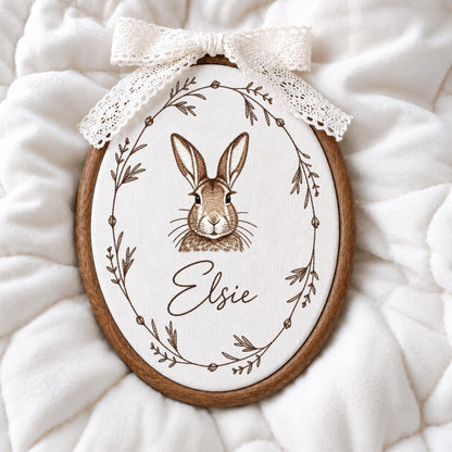 May include: An oval wooden embroidery hoop containing white fabric with a brown line drawing of a rabbit centered inside a botanical wreath. The name Elsie is printed in brown script below the rabbit. A white lace bow is attached to the top.