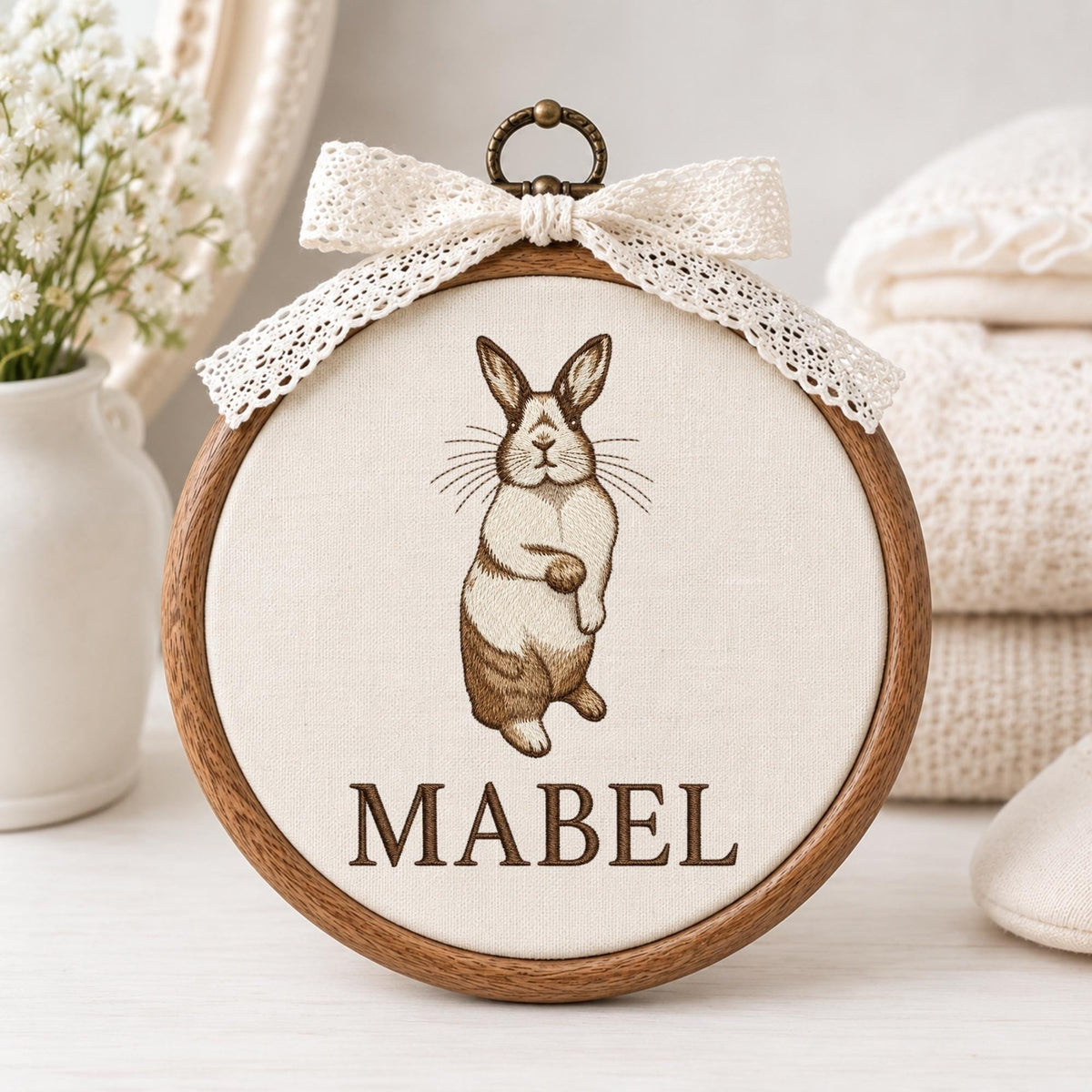 May include: A circular wooden embroidery hoop containing off-white fabric with a brown embroidered rabbit illustration and the name MABEL in brown text. A cream-colored lace ribbon is tied in a bow at the top metal hanging loop of the frame.