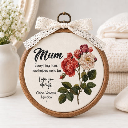May include: A circular wooden embroidery hoop containing off-white fabric with a printed floral design of red and white roses. The text reads: Mum, Everything I am, you helped me to be. Love you always, Chloe, Vanessa &amp; Jordan x. A cream lace bow is attached.
