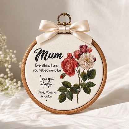 May include: A circular wooden embroidery hoop containing cream fabric with a floral design of red and white roses. The text reads: Mum, Everything I am, you helped me to be. Love you always, Chloe, Vanessa &amp; Jordan x. A cream satin bow sits at the top.
