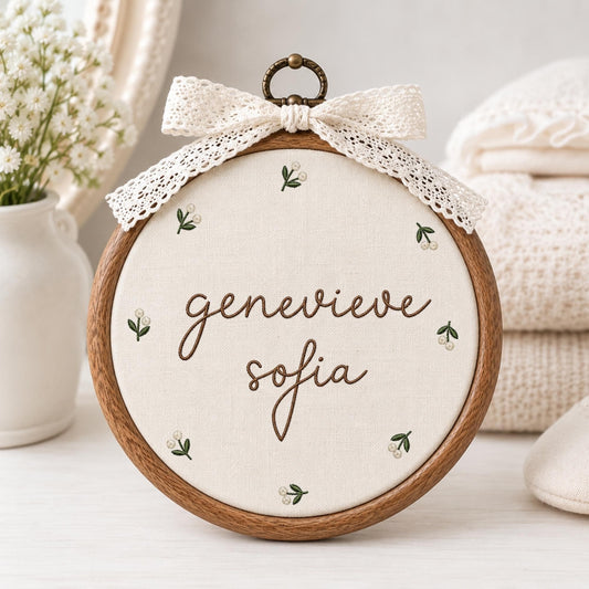 May include: A circular wooden embroidery hoop containing off-white fabric with the brown embroidered name genevieve sofia. The fabric is decorated with small green and white floral sprigs. A cream-colored lace bow is attached to the top.