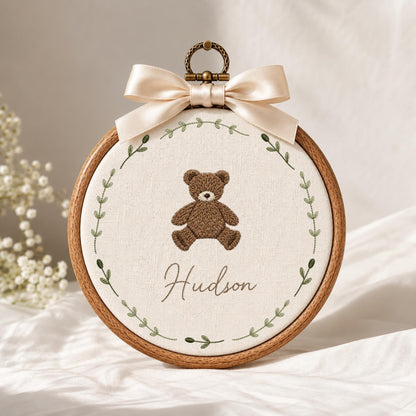 May include: A circular wooden embroidery hoop containing off-white fabric with a brown teddy bear embroidered in the center. Below the bear, the name Hudson is stitched in cursive. A green leaf wreath surrounds the design, topped with a cream ribbon.