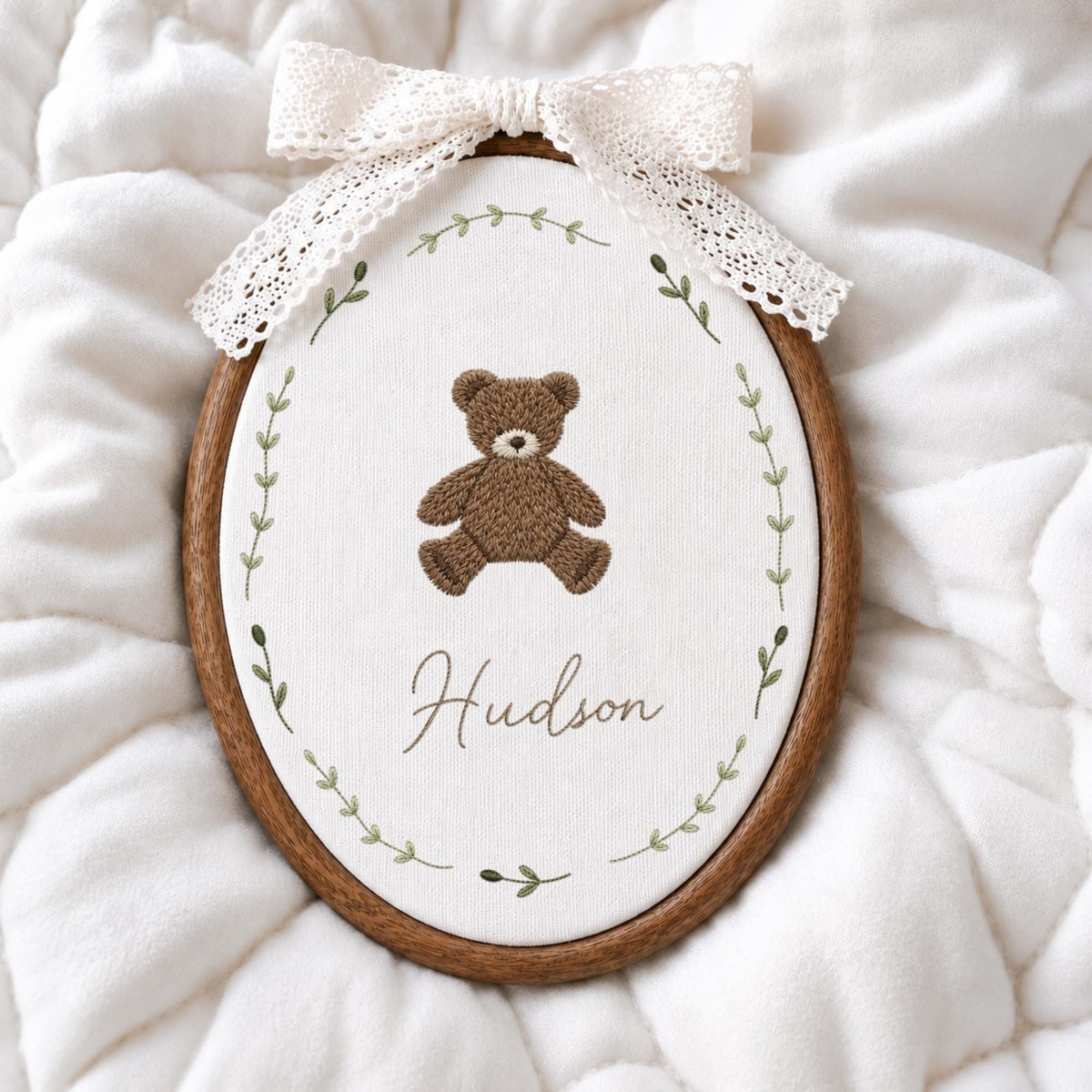 May include: An oval wooden embroidery hoop containing white fabric with a brown teddy bear stitched in the center. Below the bear, the name Hudson is embroidered in brown script. A green leaf vine encircles the design, and a white lace bow is attached to the top of the hoop.