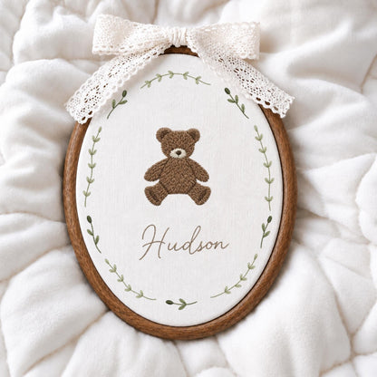 May include: An oval wooden embroidery hoop containing white fabric with a brown teddy bear stitched in the center. Below the bear, the name Hudson is embroidered in brown script. A green leaf vine encircles the design, and a white lace bow is attached to the top of the hoop.